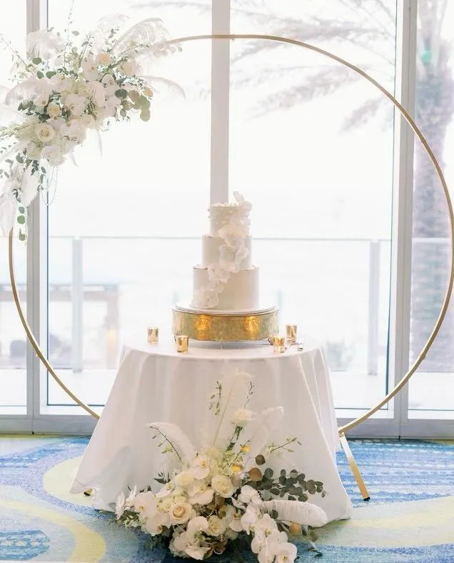 White wedding cake under a gold floral arch by the water