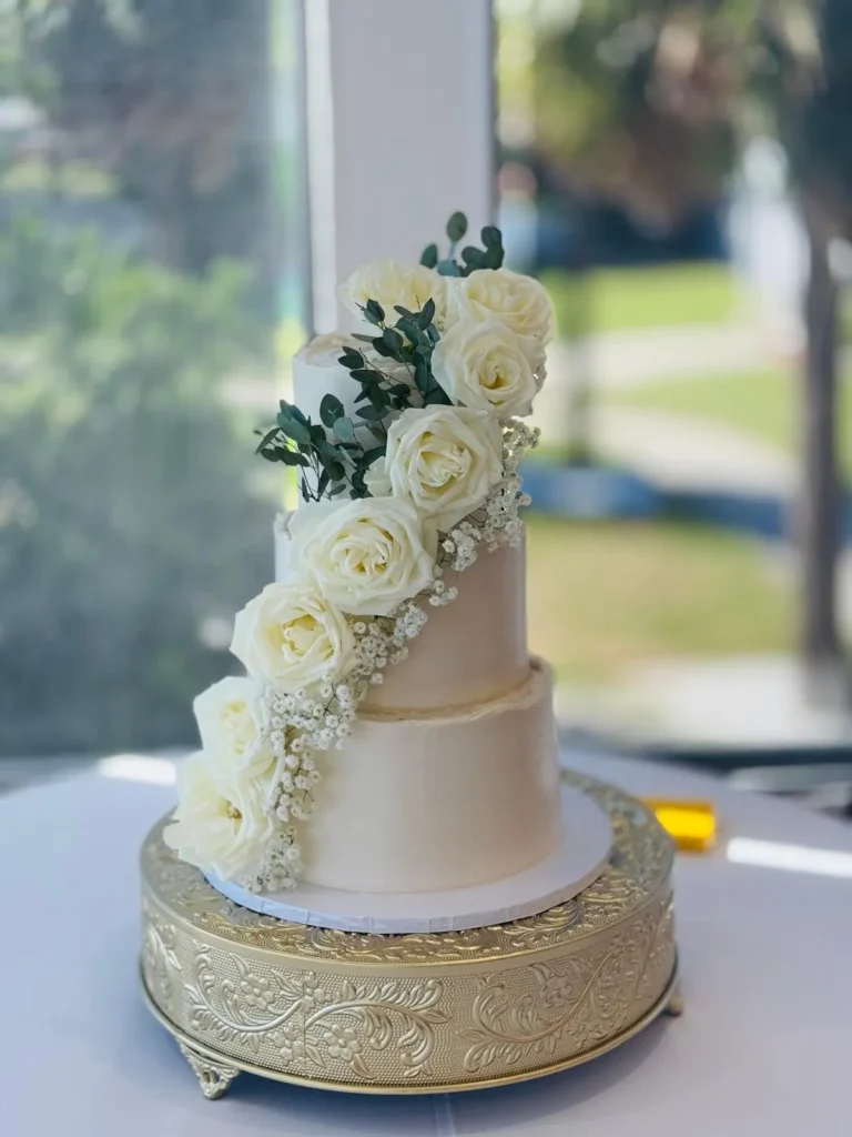 Three-tier ivory wedding cake with a white rose cascade on a gold stand