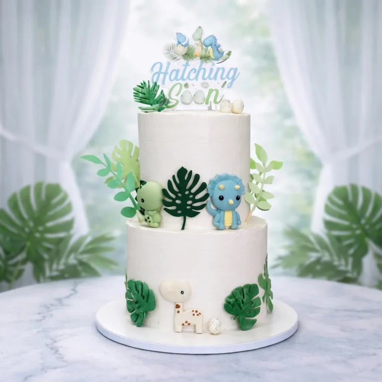 Two-tier white baby shower cake with baby dinosaurs, green leaves and a "Hatching Soon" topper
